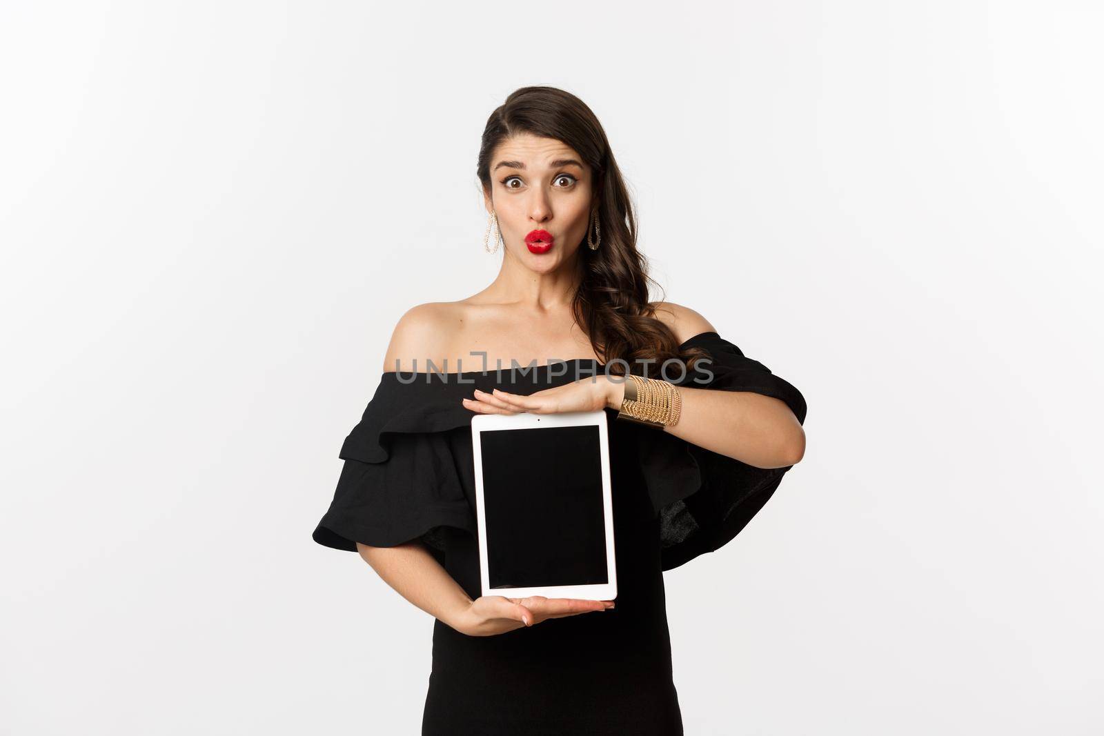 Fashion and shopping concept. Beautiful woman with red lipsticks, black dress, showing tablet screen and looking excited, standing over white background.