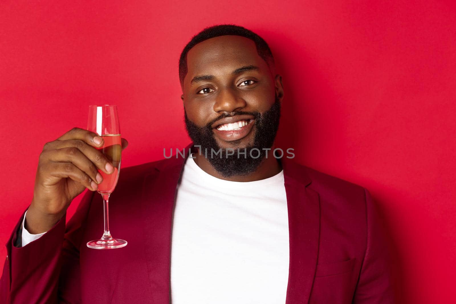 Close-up of handsome smiling Black man saying toast, raising glass of champagne for merry christmas and happy new year, celebrating against red background.