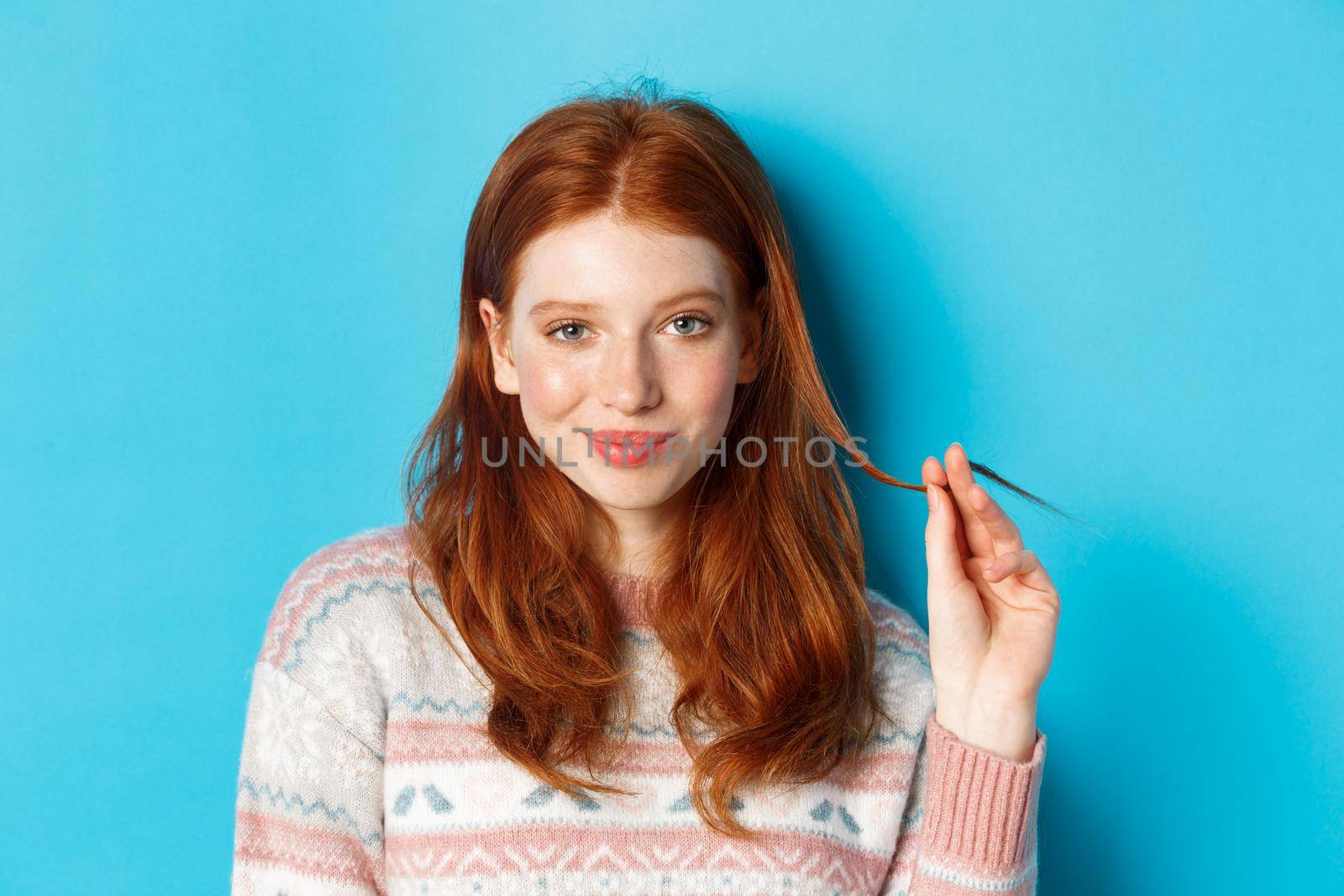 Close-up of confident and sassy redhead teen girl looking at camera pleased, playing with hair strand and smirking, standing over blue background.