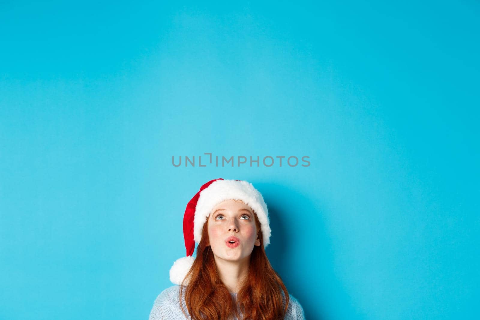 Winter holidays and Christmas eve concept. Head of pretty redhead girl in santa hat, appear from bottom and looking up at logo impressed, seeing promo offer, blue background by Benzoix