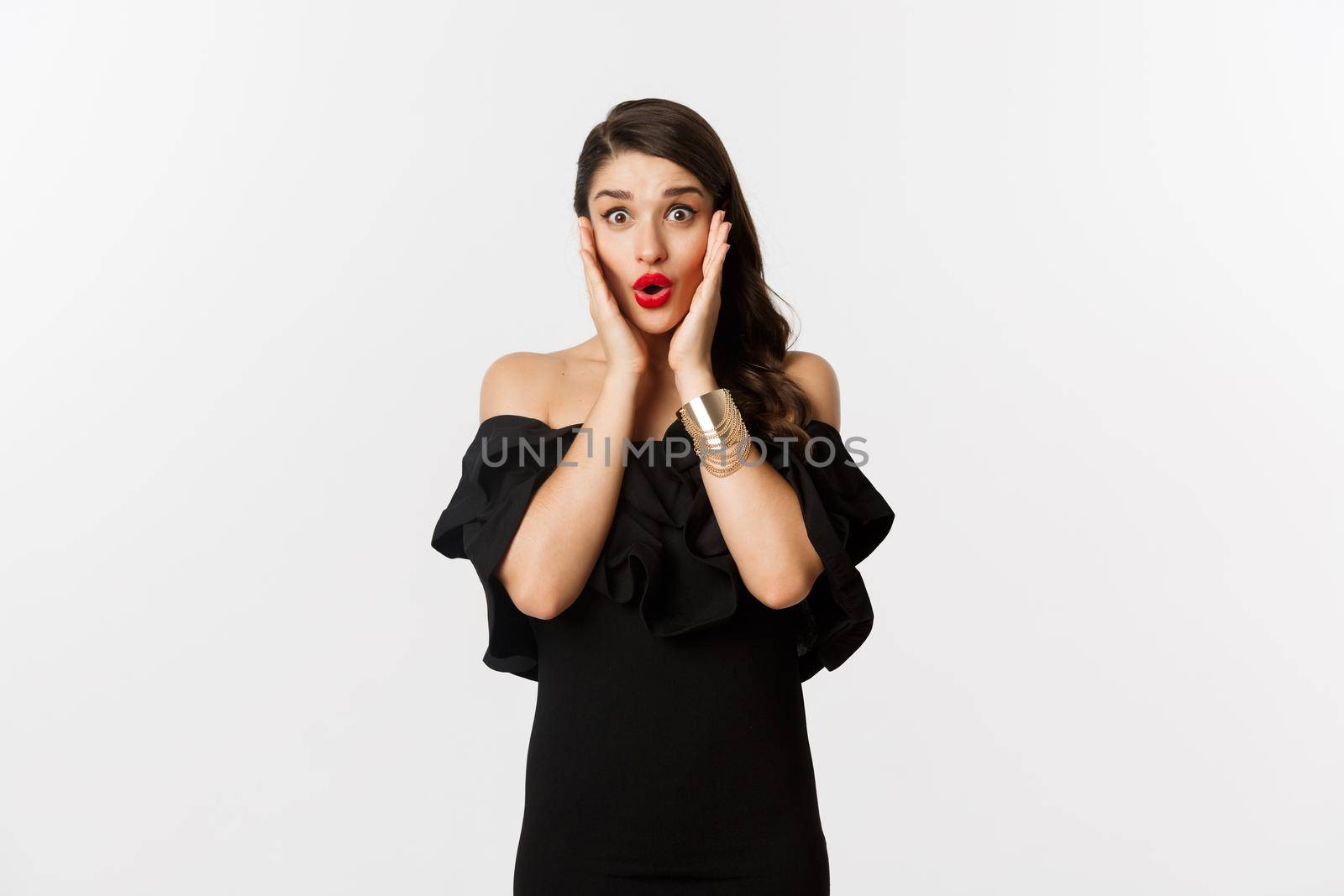 Beauty and fashion concept. Attractive woman in glamour black dress, jewelry and makeup, looking surprised and excited, standing over white background.
