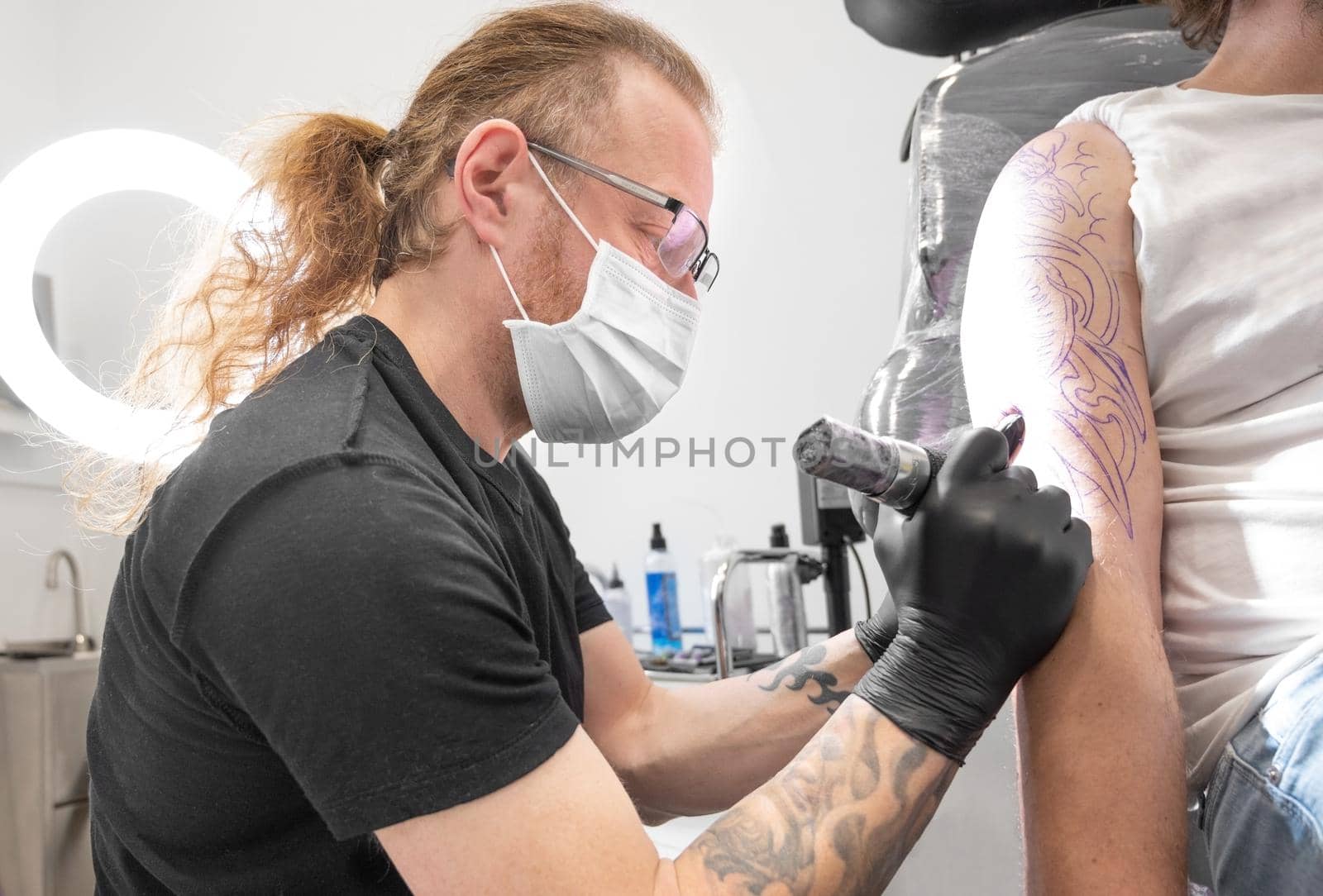 Tattooist with his machine tattooing on the arm with the design drawn and using protective mask against covid 19, concept of art and design during the new normal. by HERRAEZ