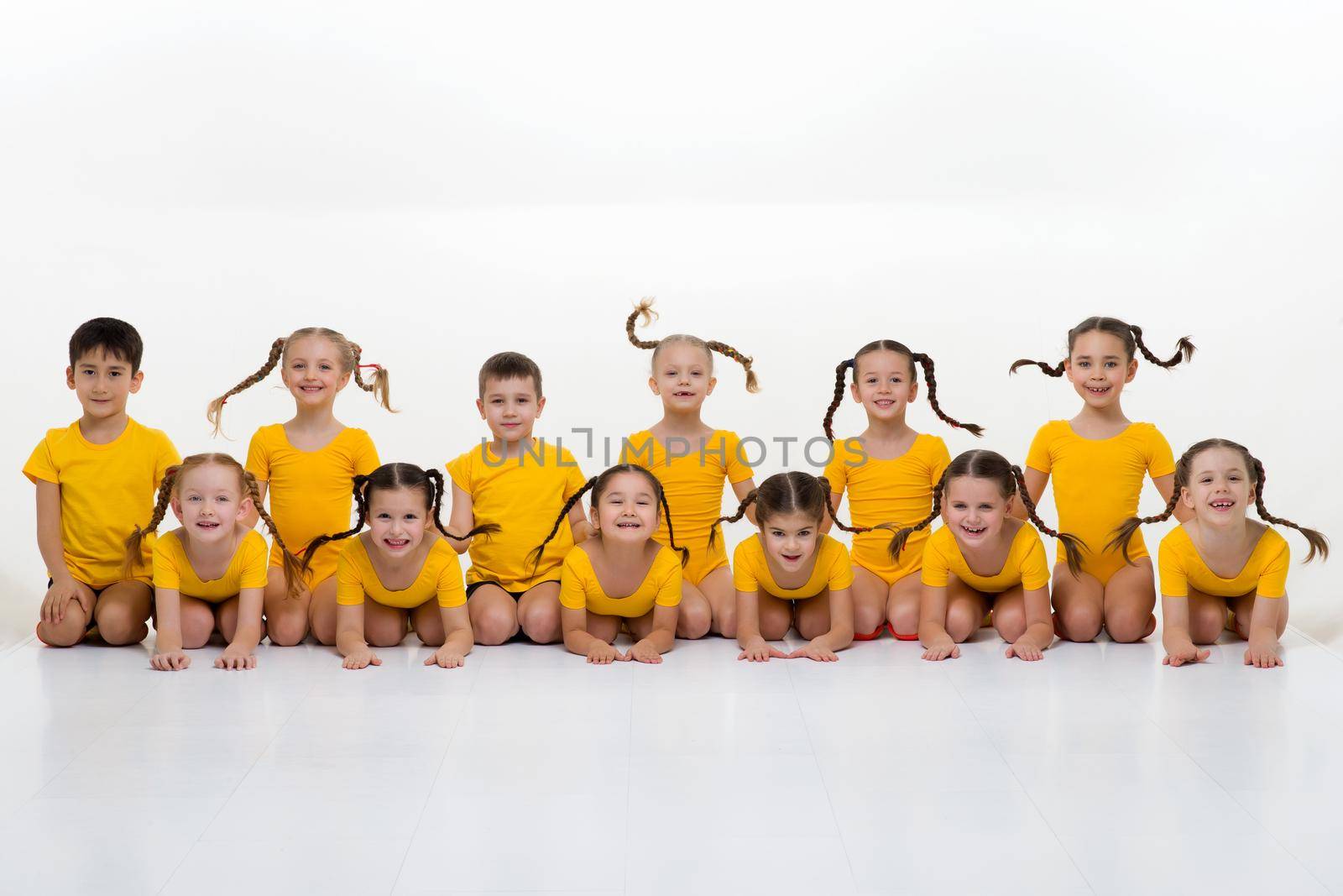 Dancer team sitting on floor in row posing in studio. Group of happy little boys and girls in yellow sportswear posing against white background. Dance school, kids education concept