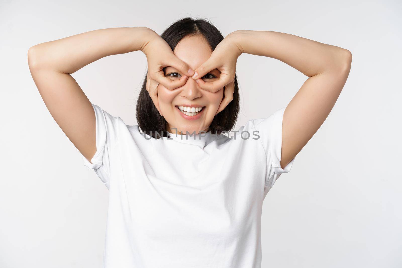 Funny young asian woman, korean girl making eyes glasses gesture, looking happy at camera, standing over white background.