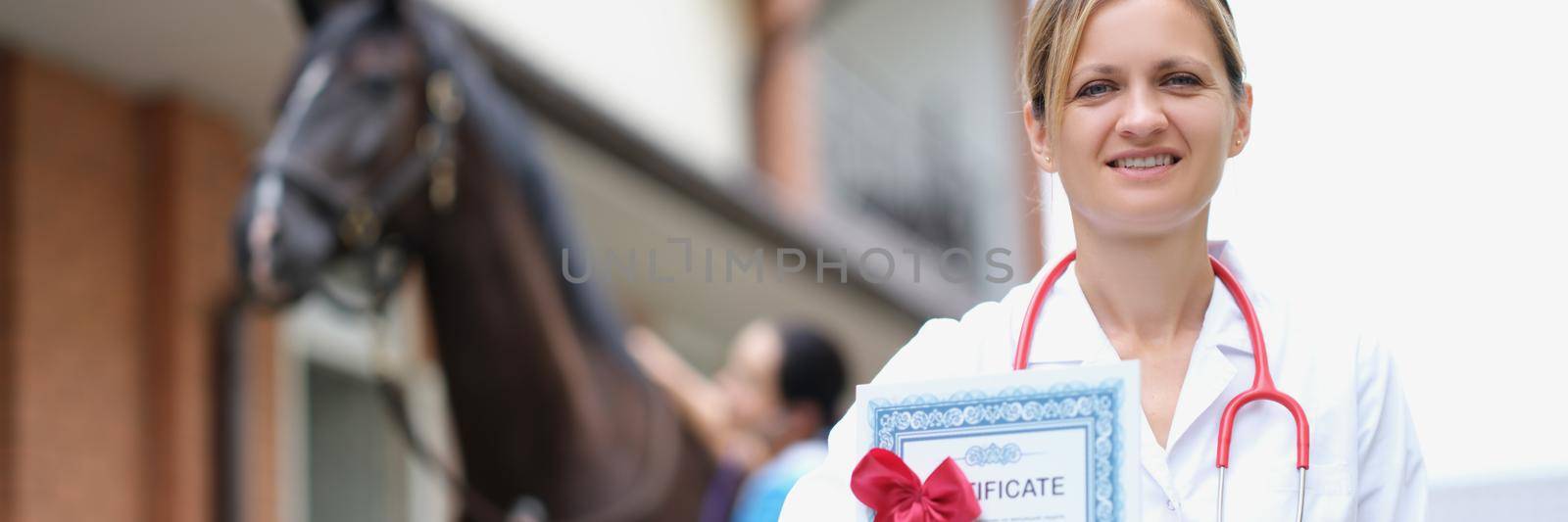 Portrait of smiling female veterinarian holding medical certificate against background of sport horse. Horses undergoing medical examination before competition concept