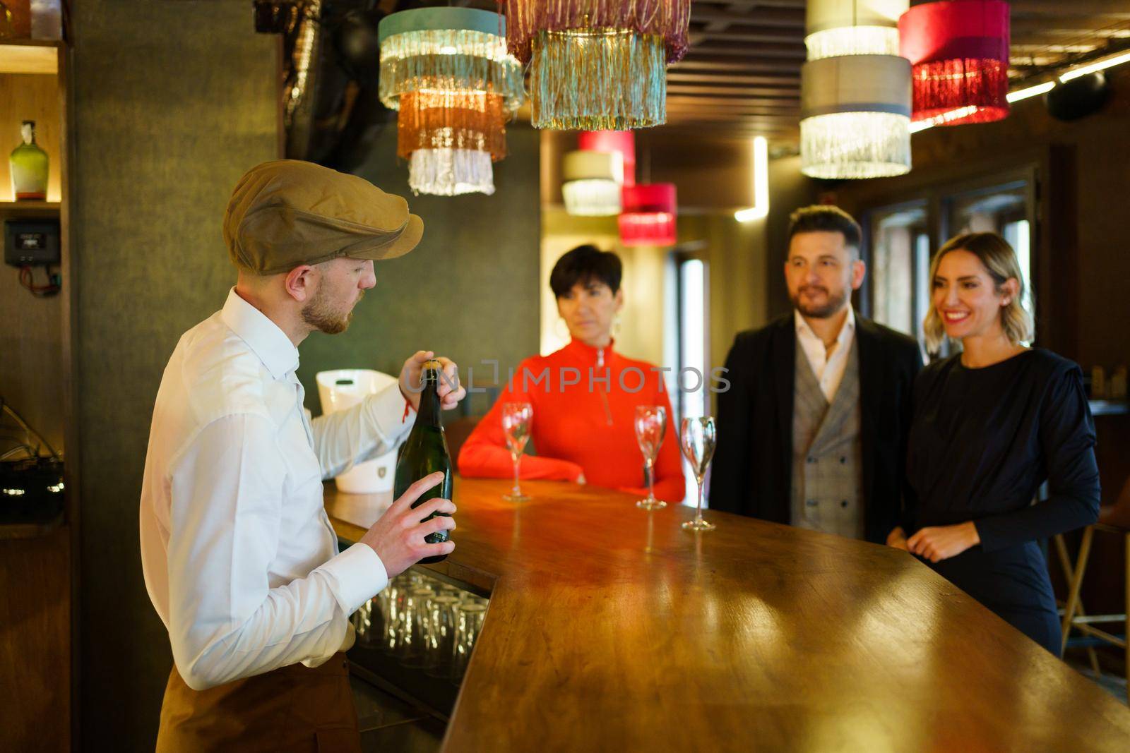 Side view of barman opening bottle of champagne and serving group of elegant people standing at wooden counter