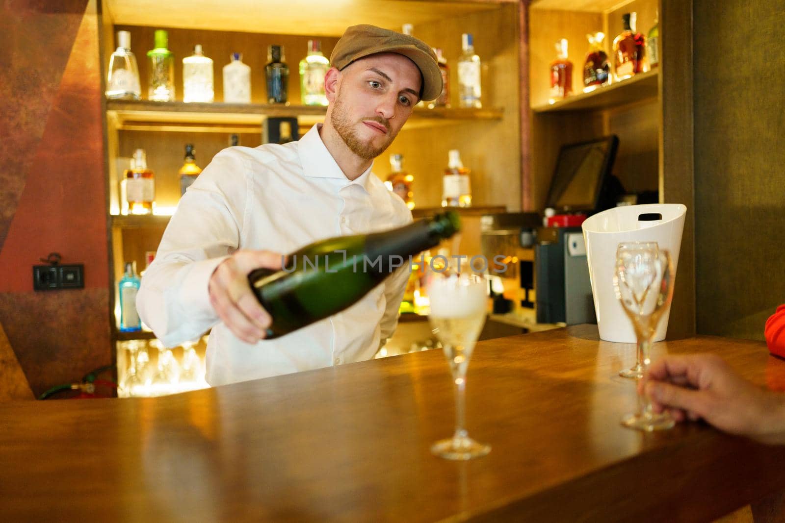 Elegant male bartender pouring sparkling champagne in glasses placed on wooden counter in luxury bar with crop unrecognizable client