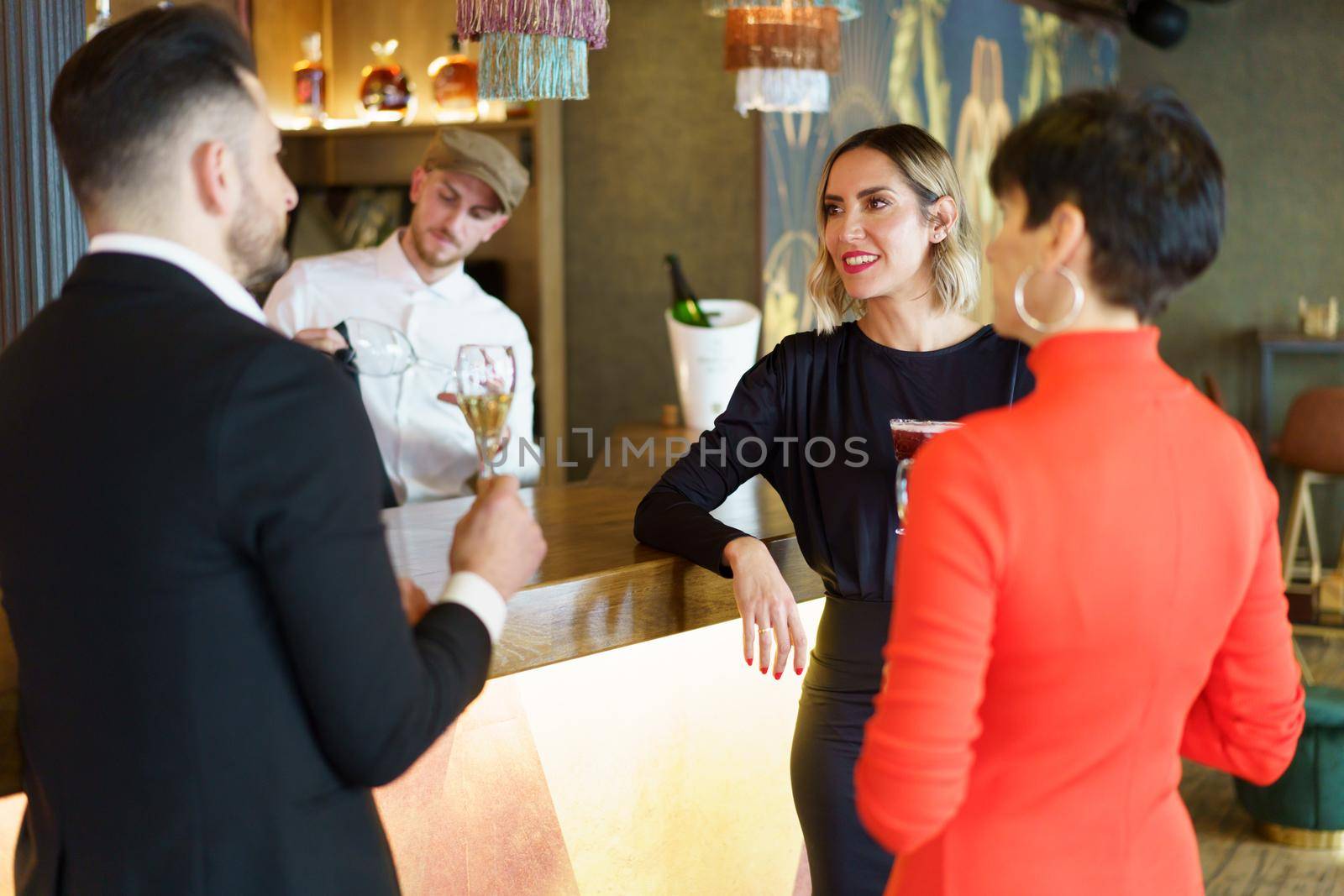 Group of well dressed friends drinking alcohol beverages and enjoying meeting while standing at wooden counter in bar with bartender