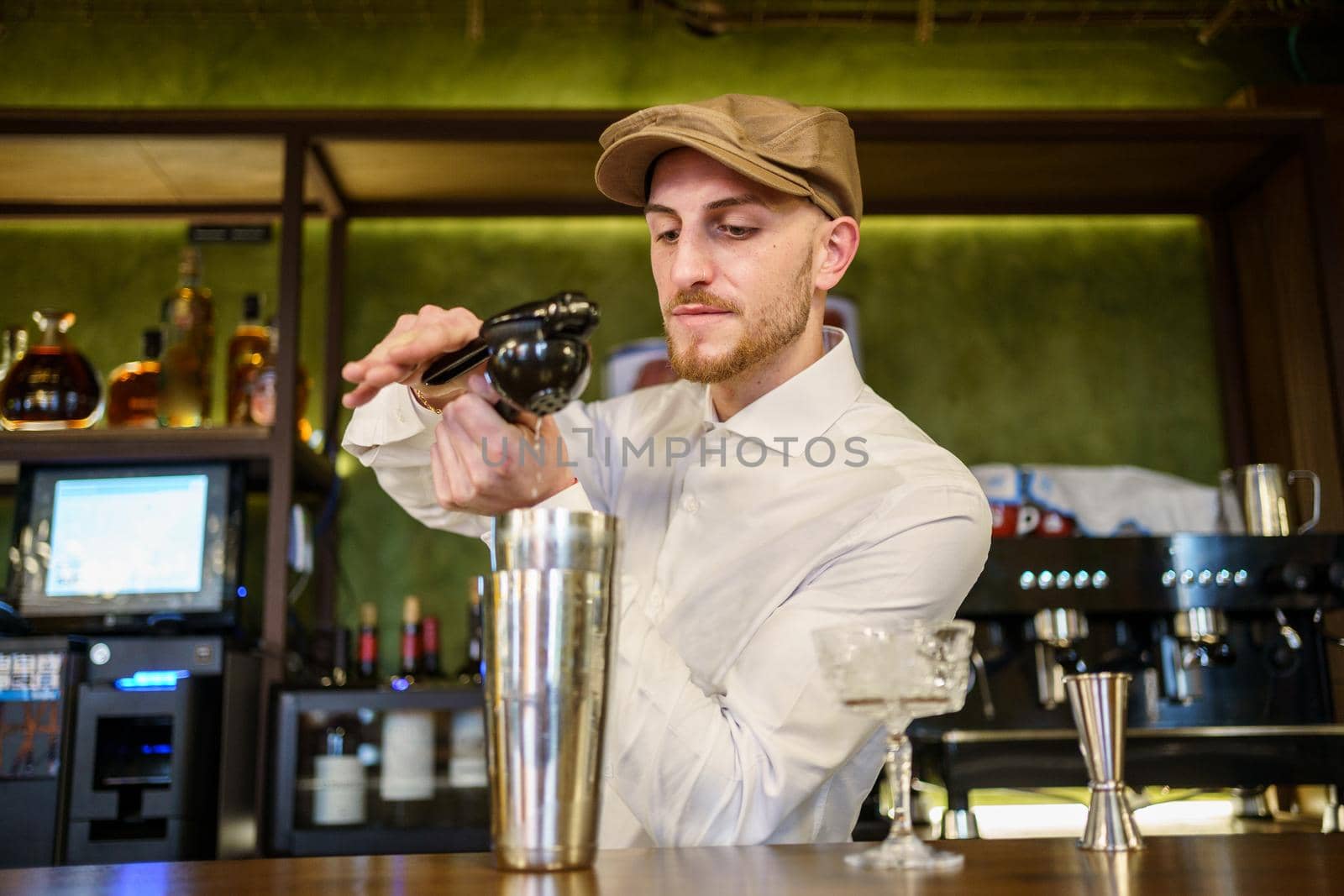 Bearded man in white shirt and cap squeezing lemon juice over shaker with cocktail while working behind counter in pub