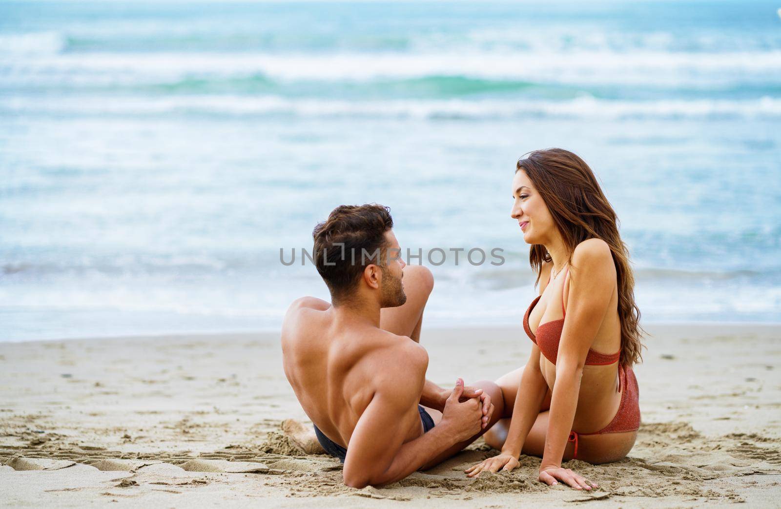 Cheerful loving woman in bikini and man in shorts sitting on shore in summer and looking at each other