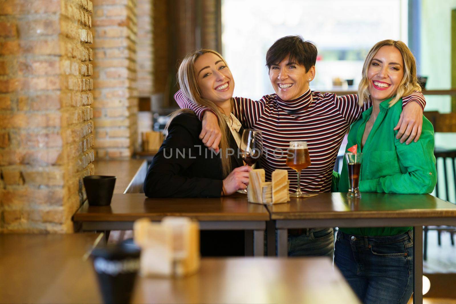 Cheerful group of female friends looking at camera with smile and embracing girlfriends behind table with alcohol during meeting in pub