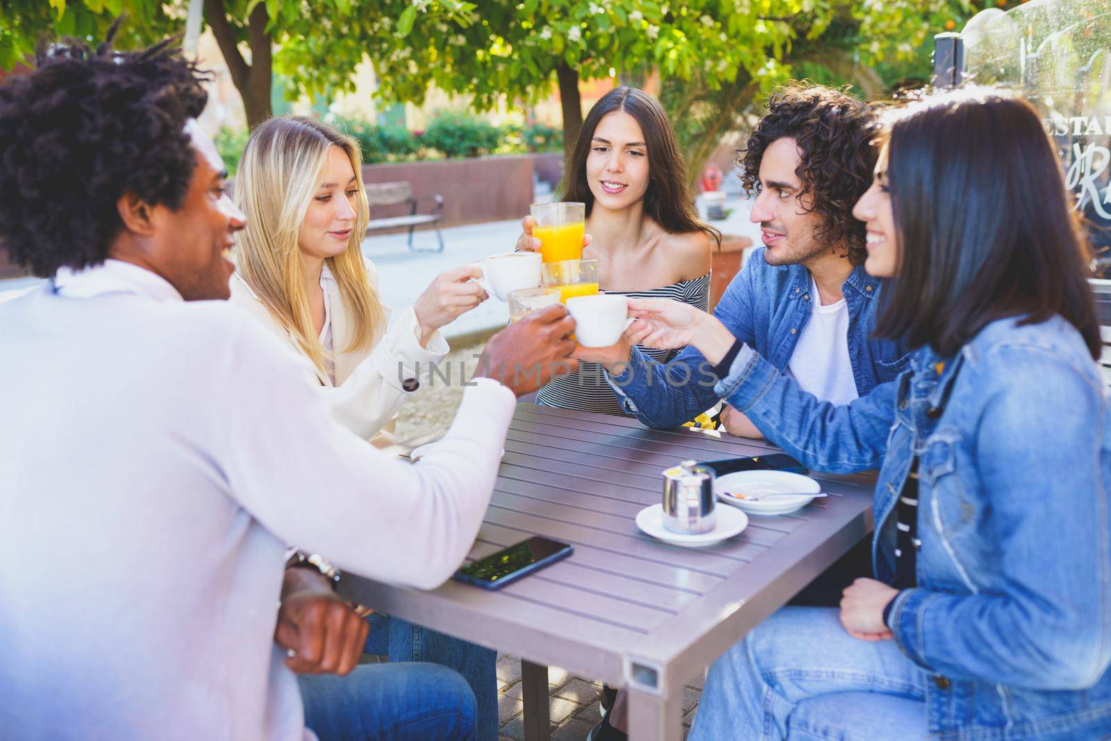 Group of happy diverse friends clinking cups and glasses while gathering at table in outdoors cafe and celebrating meeting in summer
