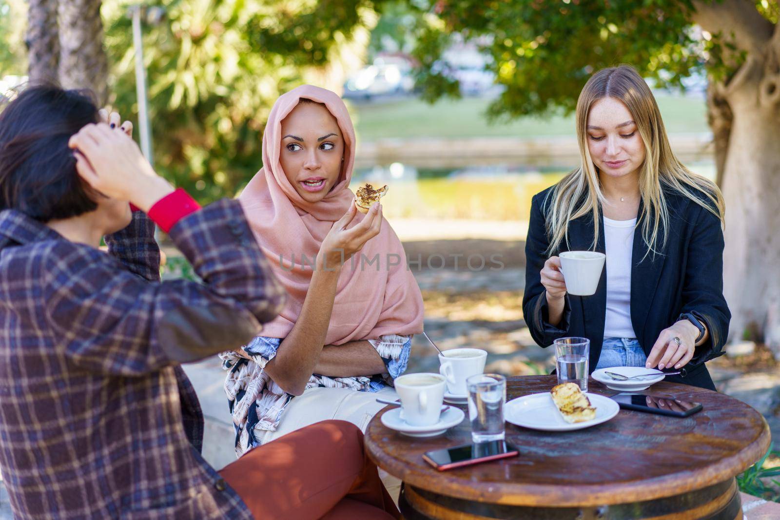Group of diverse female friends chatting while sitting at table with food and cups of coffee on terrace of outdoor cafeteria