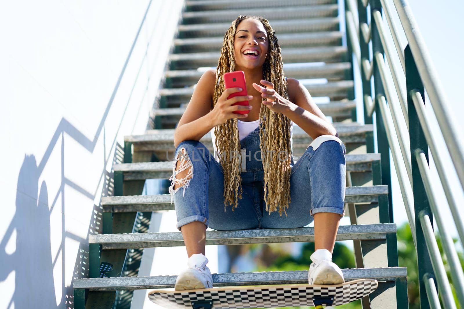 Black girl with coloured braids, sitting on some steps with a smartphone and a skateboard.