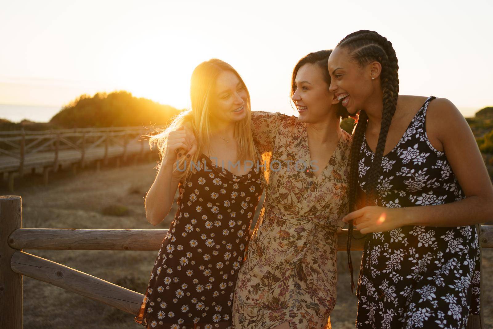 Smiling multiracial female friends hugging and looking at each other while standing near wooden fencing on embankment with bright sunlight