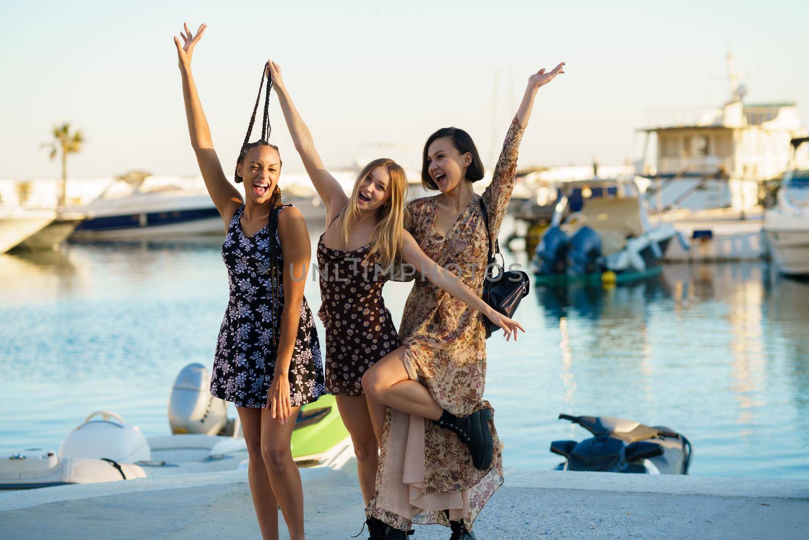 Cheerful multiracial female friends looking at camera with raised arms while standing in port near river with boats on summer day