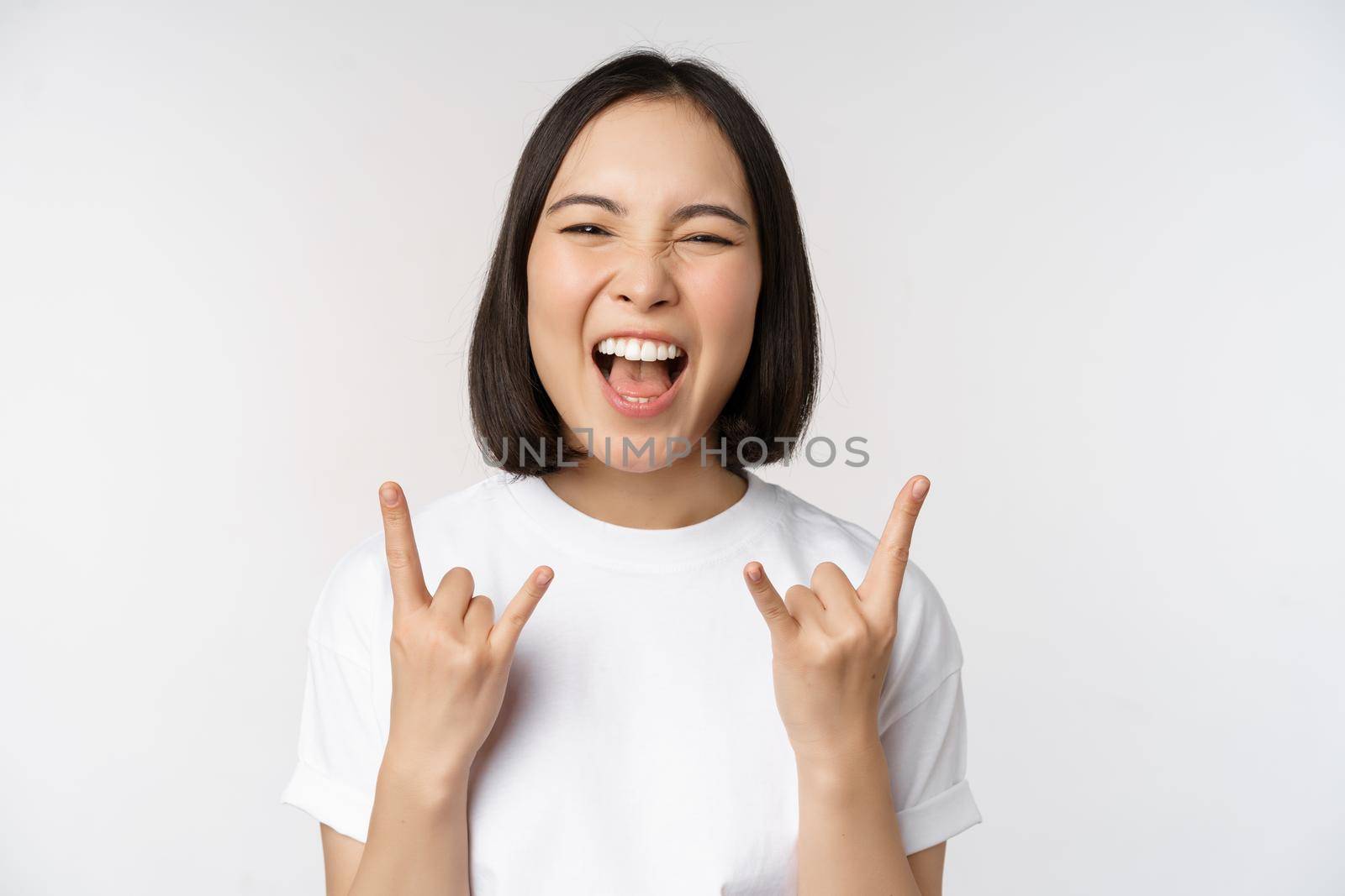 Sassy asian girl shouting, enjoying concert or festival, showing rock on, heavy metal sign, having fun, standing over white background.