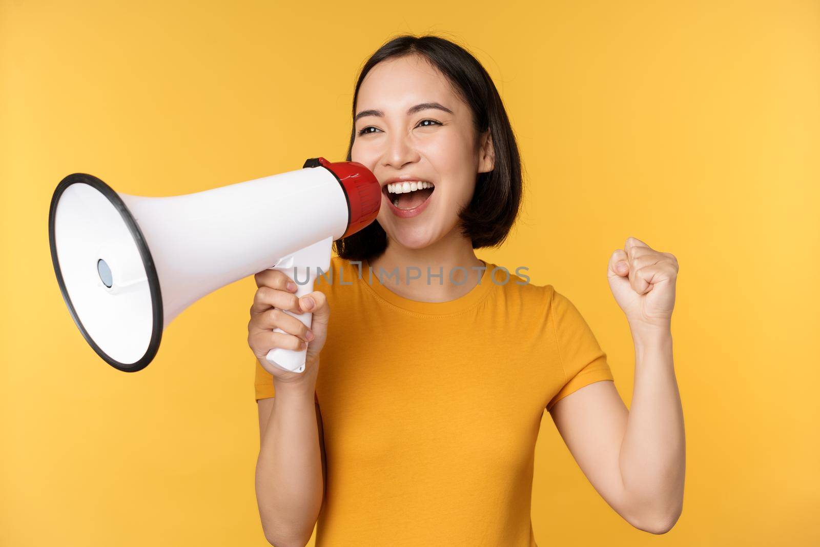 Announcement. Happy asian woman shouting loud at megaphone, recruiting, protesting with speaker in hands, standing over yellow background.