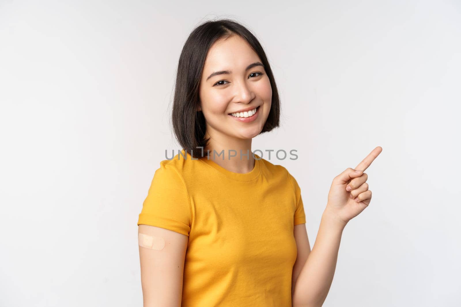 Covid-19 and vaccination concept. Smiling asian girl with band aid on shoudler, pointing finger at banner, showing vaccine campaign, standing over white background by Benzoix