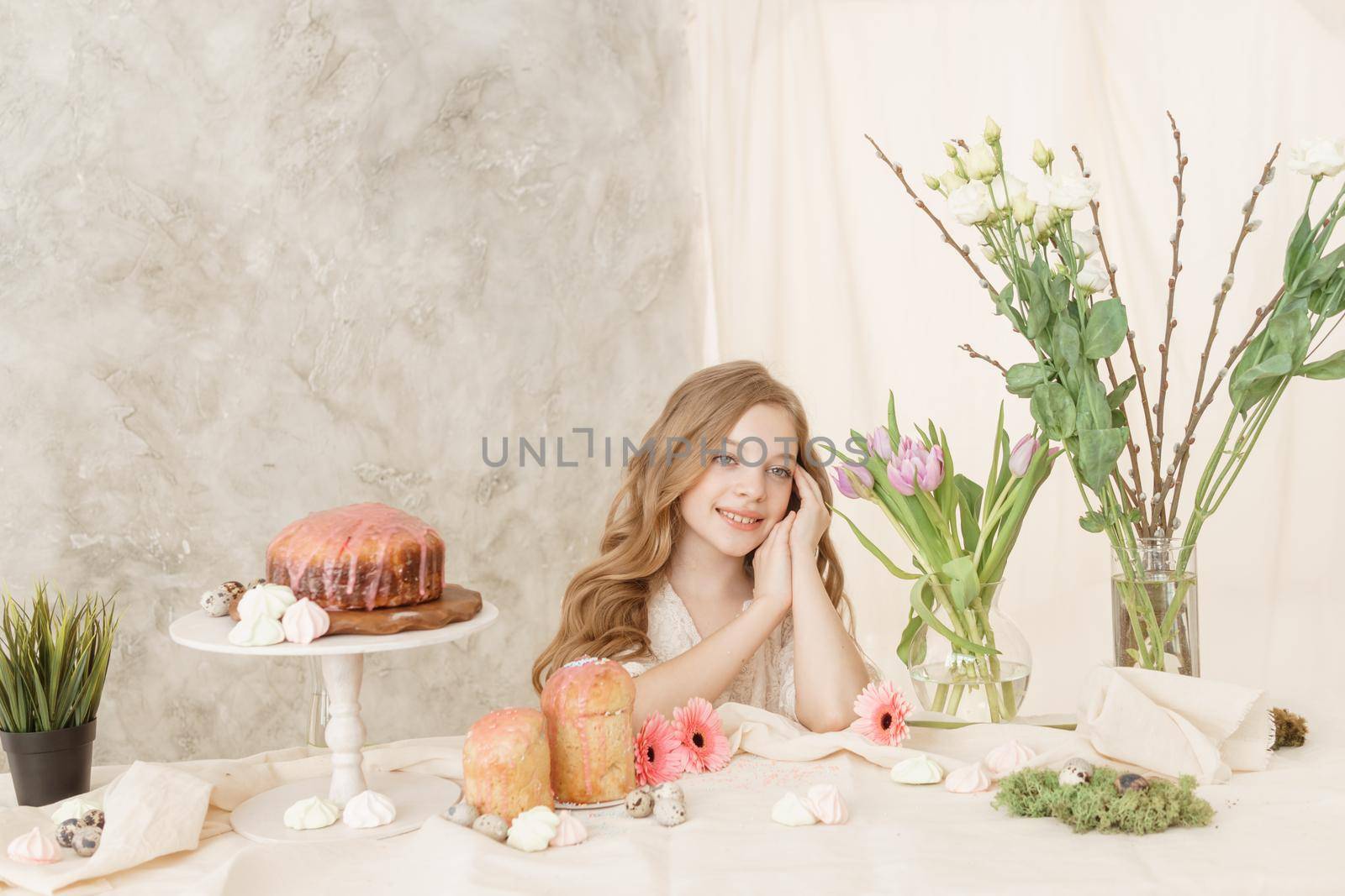 A girl with long hair in a light dress is sitting at the Easter table with cakes, spring flowers and quail eggs. Happy Easter celebration