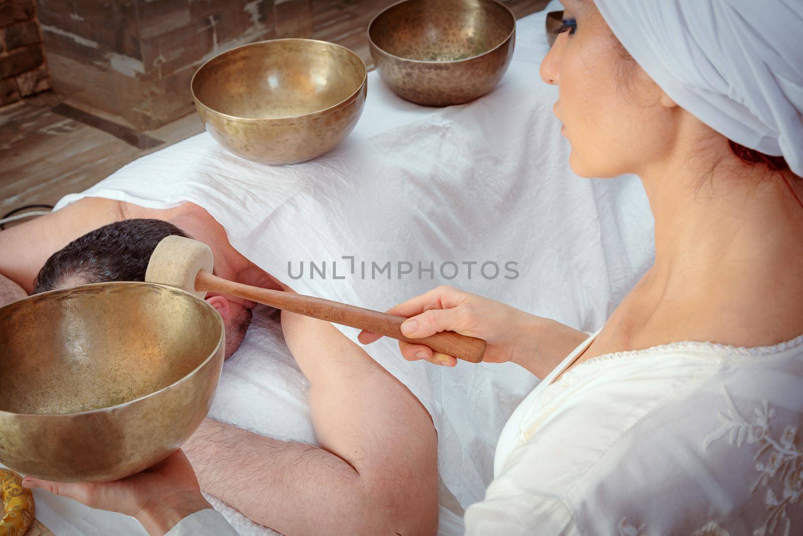 A woman performs Tibetan singing bowl therapy with a man lying under a white sheet. Relaxing meditation.