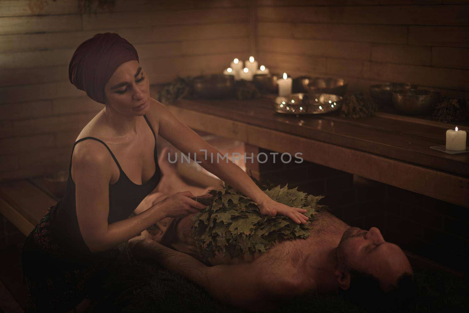 A woman conducts a bathing ritual for a man with the help of brooms. A man lies on spruce branches.