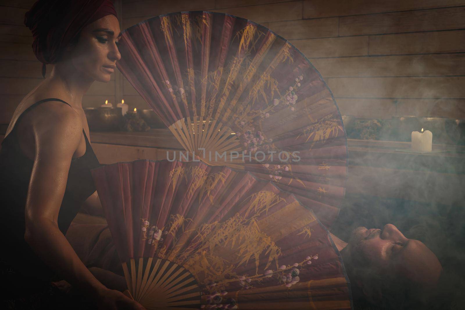 A woman performs a healing ritual with a Chinese fan for a man. A man lies on spruce branches.