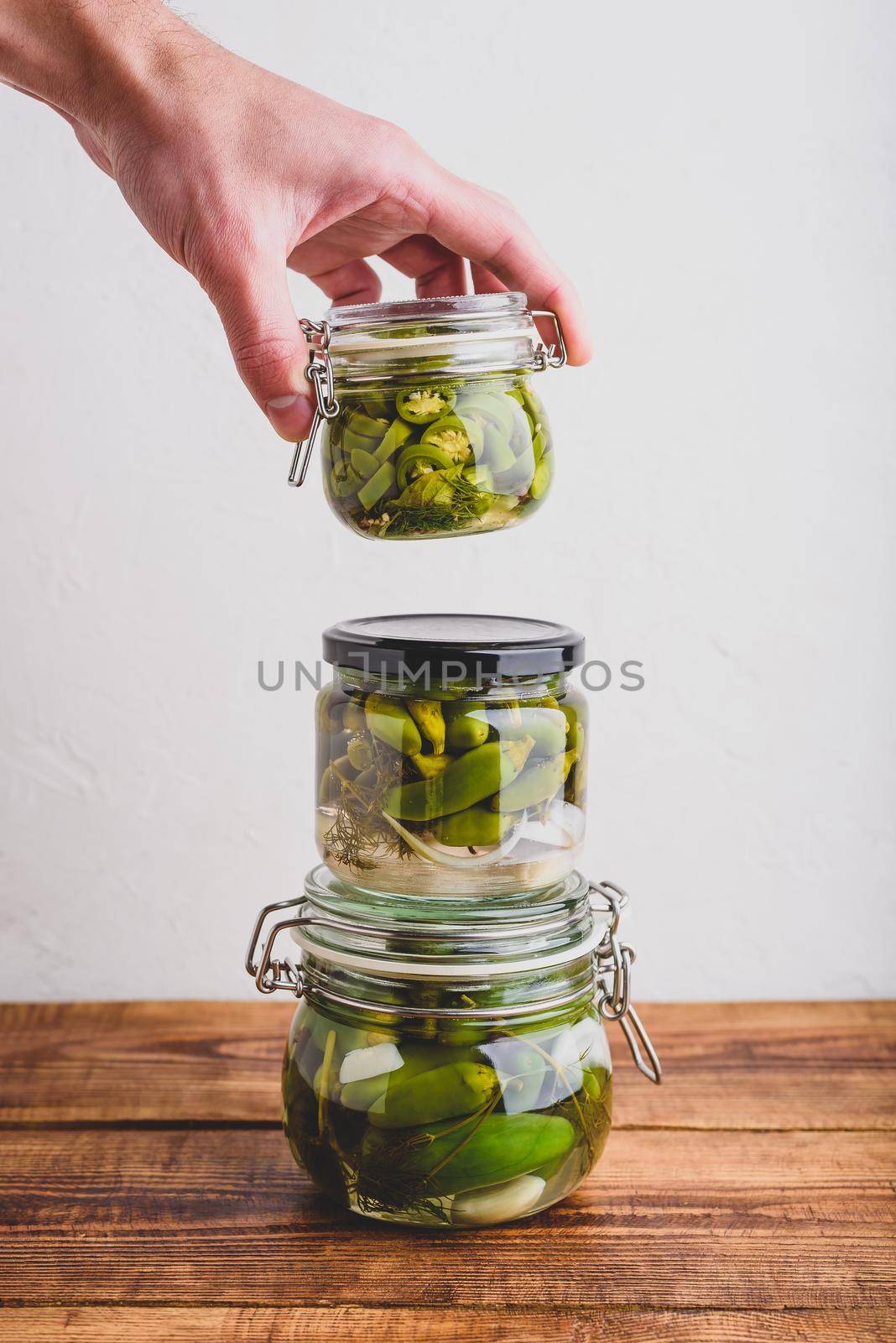 Male Hand Holding One of Three Glass Jars of Freshly Pickled Jalapeno Peppers with Herbs and Garlic. Copy Space