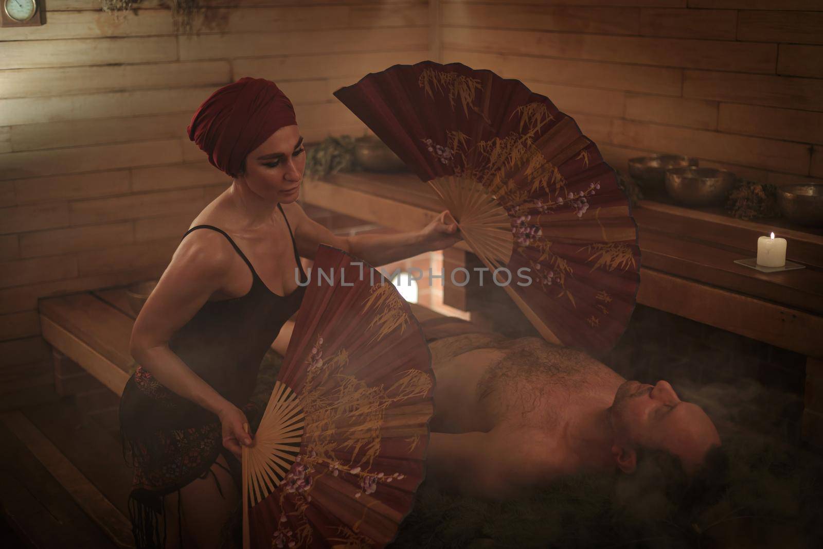 A woman performs a healing ritual with a Chinese fan for a man. A man lies on spruce branches by Yurich32