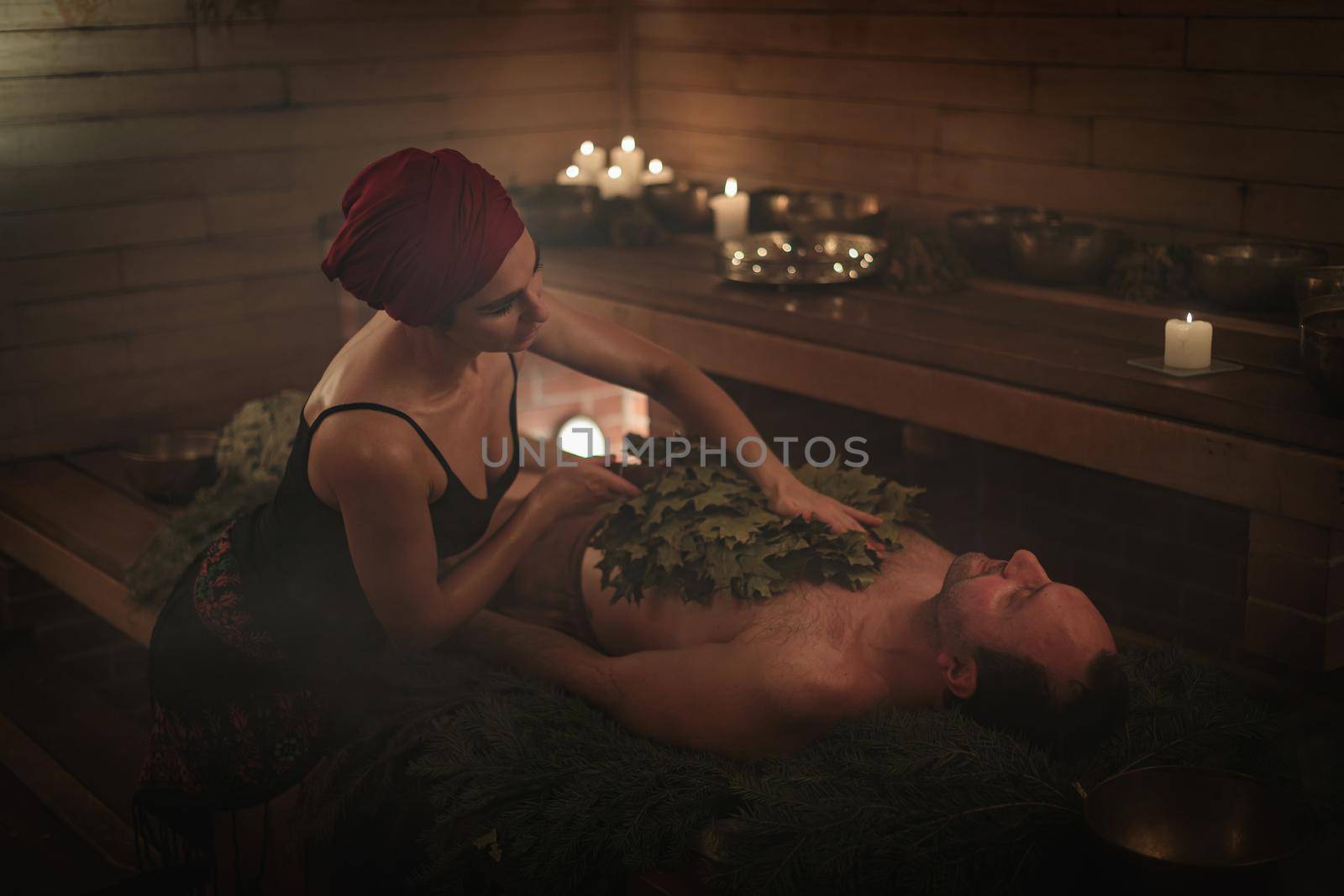 A woman conducts a bathing ritual for a man with the help of brooms. A man lies on spruce branches by Yurich32