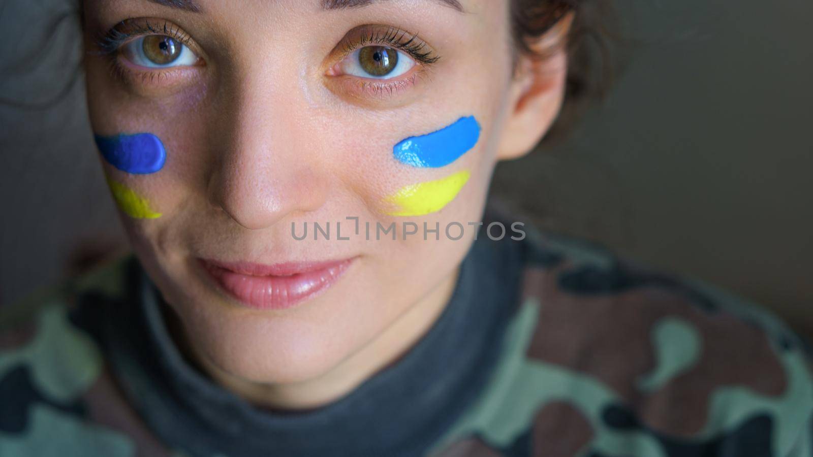 Indoor portrait of young girl with blue and yellow ukrainian flag on her cheek wearing military uniform, mandatory conscription in Ukraine, equality concepts.