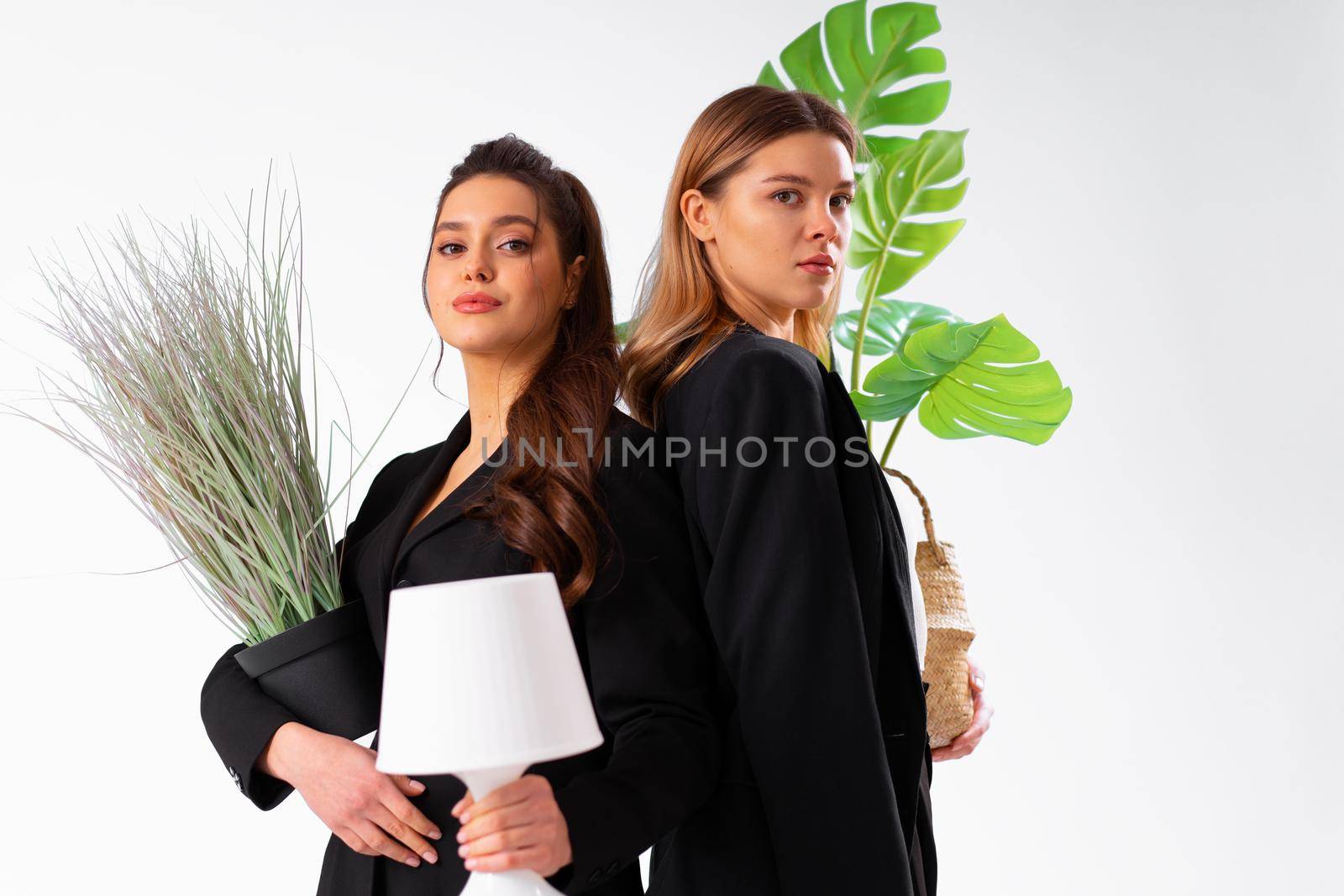 Two young business woman dressed black suit smile holding houseplant and lamp standing isolated on white background. Moving concept. Relocation new office with positive. by andreonegin