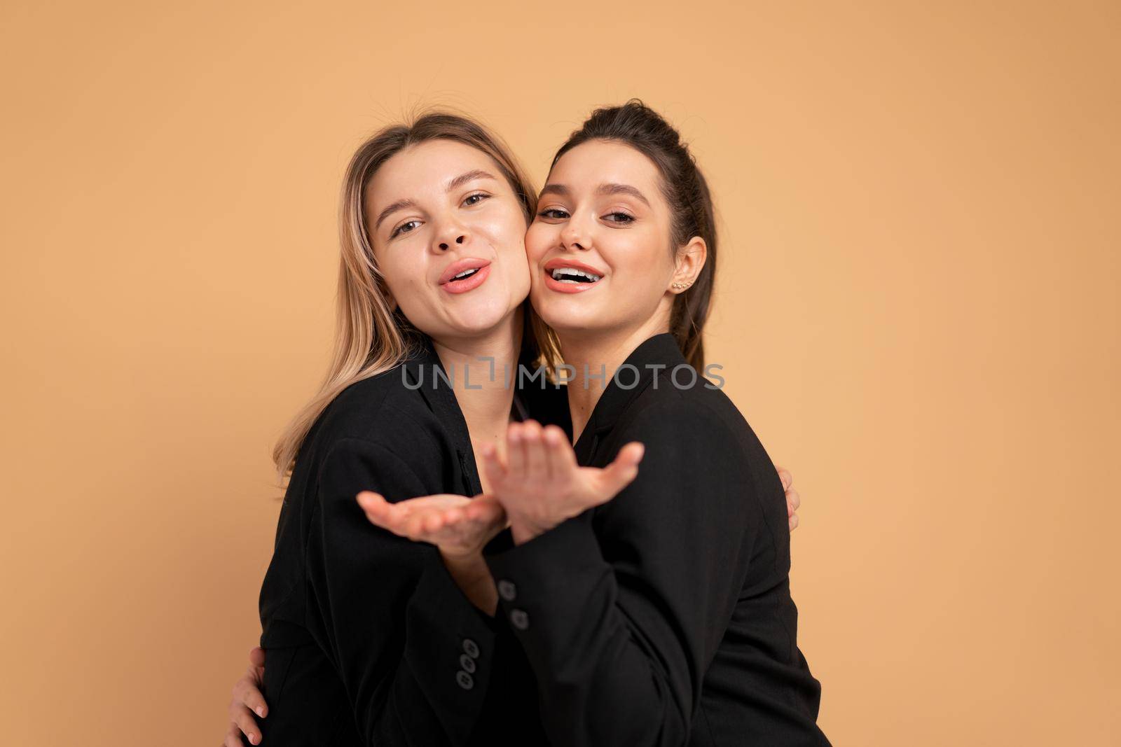 Two young business woman dressed black suit embrace laugh and happy standing in studio isolated over peach color background. Female friends and partners in business concept sending blow kiss in camera