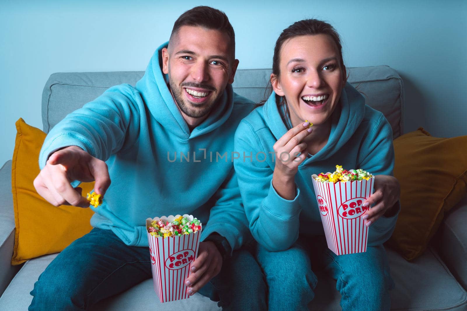 Young happy couple with popcorn sitting on sofa and watching funny comedy movie. Couple enjoying time together at home by DariaKulkova