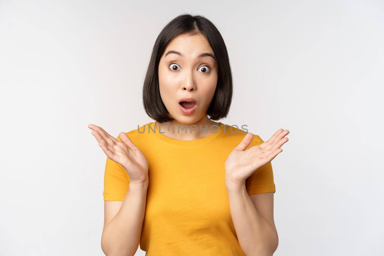 Close up portrait of asian woman looking surprised, wow face, staring impressed at camera, standing over white background in yellow t-shirt.