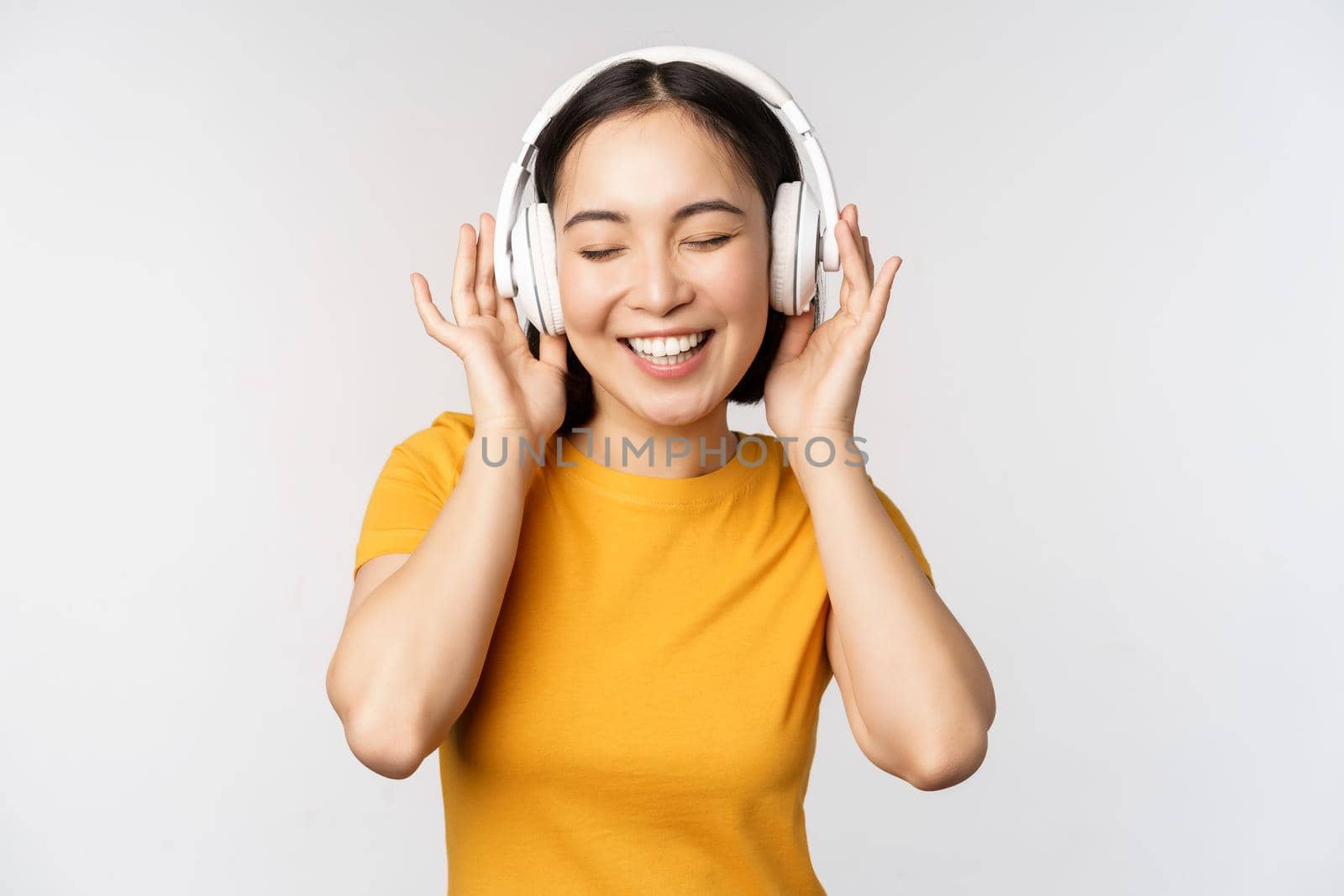 Happy asian girl dancing, listening music on headphones and smiling, standing in yellow tshirt against white background. Copy space