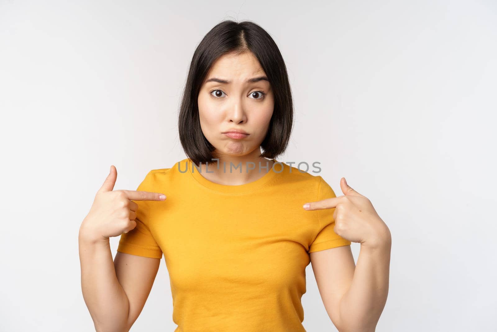 Confused asian woman pointing at herself, looking in disbelief, standing over white background by Benzoix