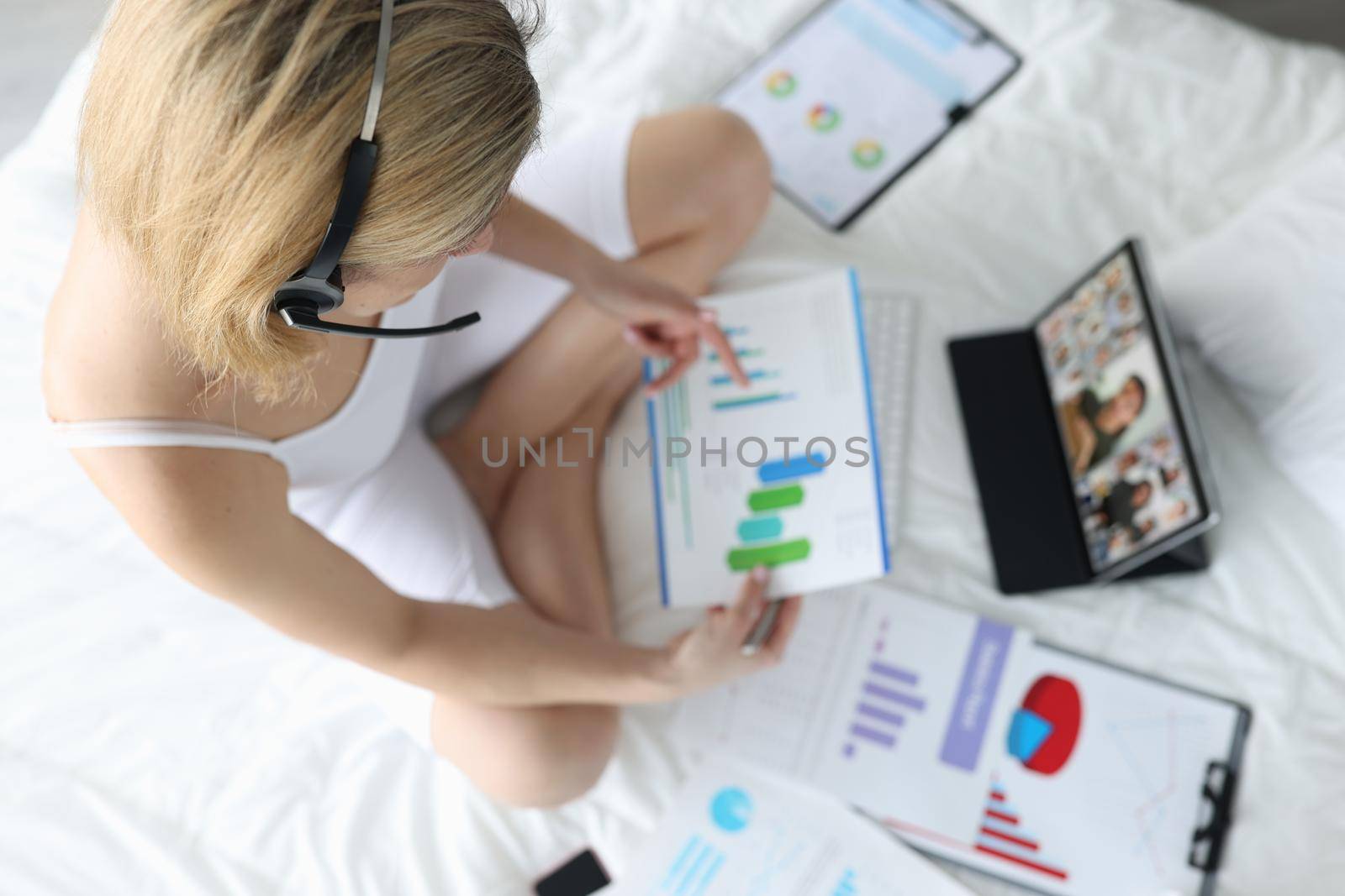 Woman with handsfree sitting on white bed with documents in hands and communicating via video link with colleagues top view by kuprevich