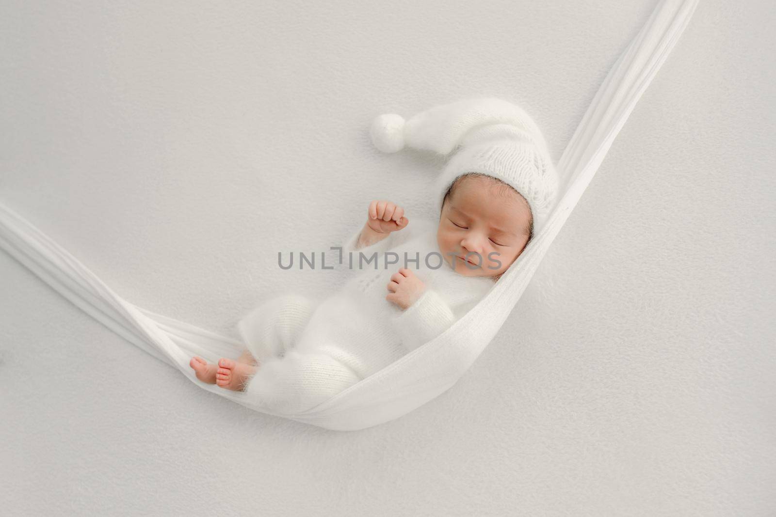 Newborn baby boy wearing white knitted costume sleeping in hammock. Infant child kid in festive holiday winter dress studio portrait