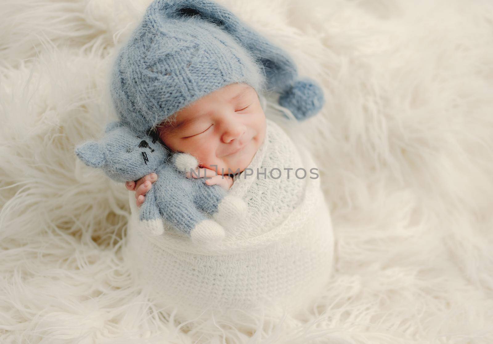 Newborn baby boy with knitted kitty cat toy sleeping and smiling swaddled in fabric on white fur. Infant child kid napping Christmas and New Year holiday portrait