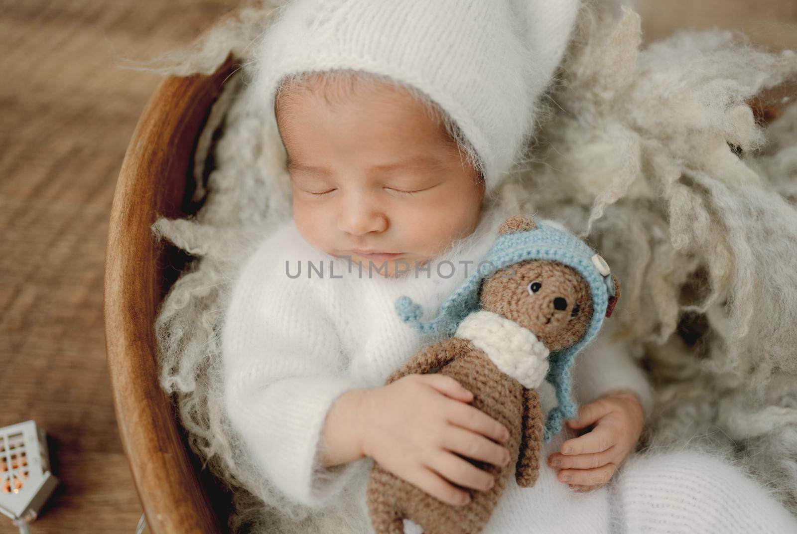 Newborn baby boy wearing white costume holding knitted teddy bear toy and sleeping on fur. Infant child kid napping studio portrait