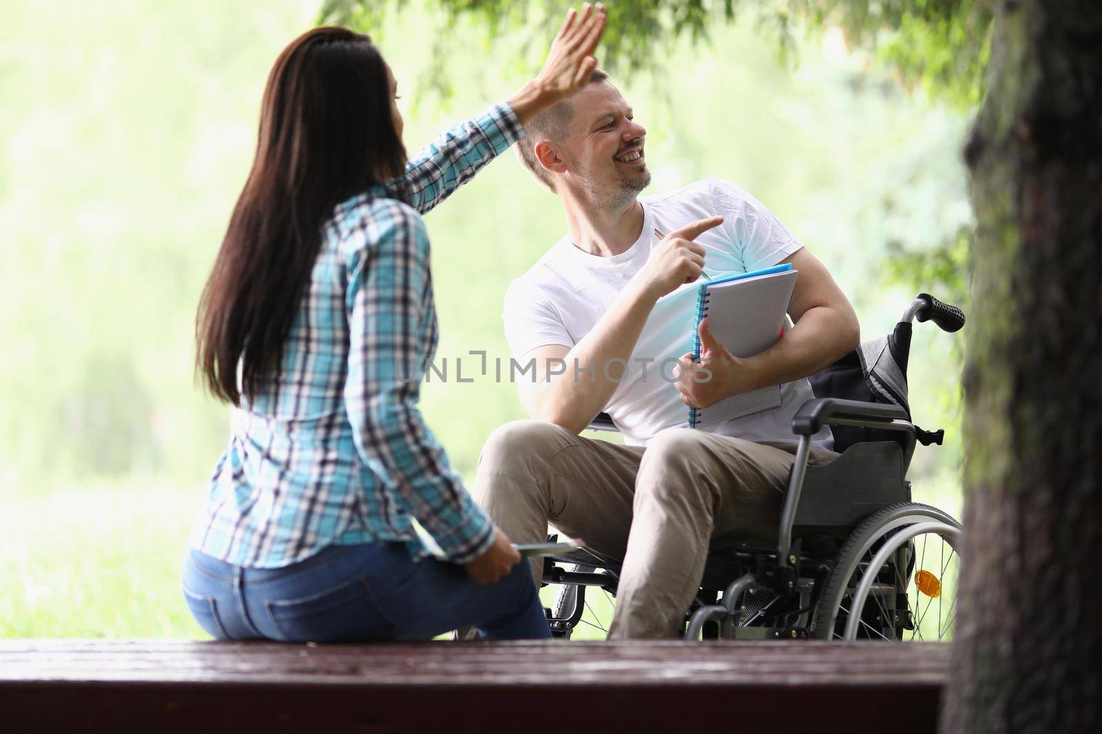 Portrait of woman and man in wheelchair wave hello to someone, people study in park, guy after accident get treatment. Leisure, recovery, nature concept