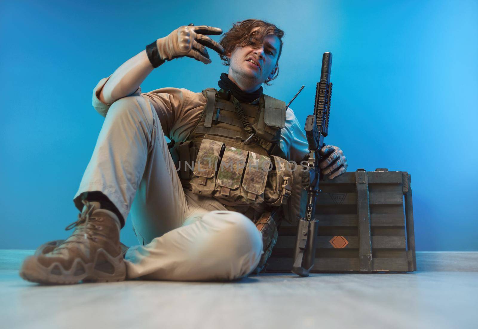 male soldier in camouflage is sitting by an ammunition crate on the floor with a weapon