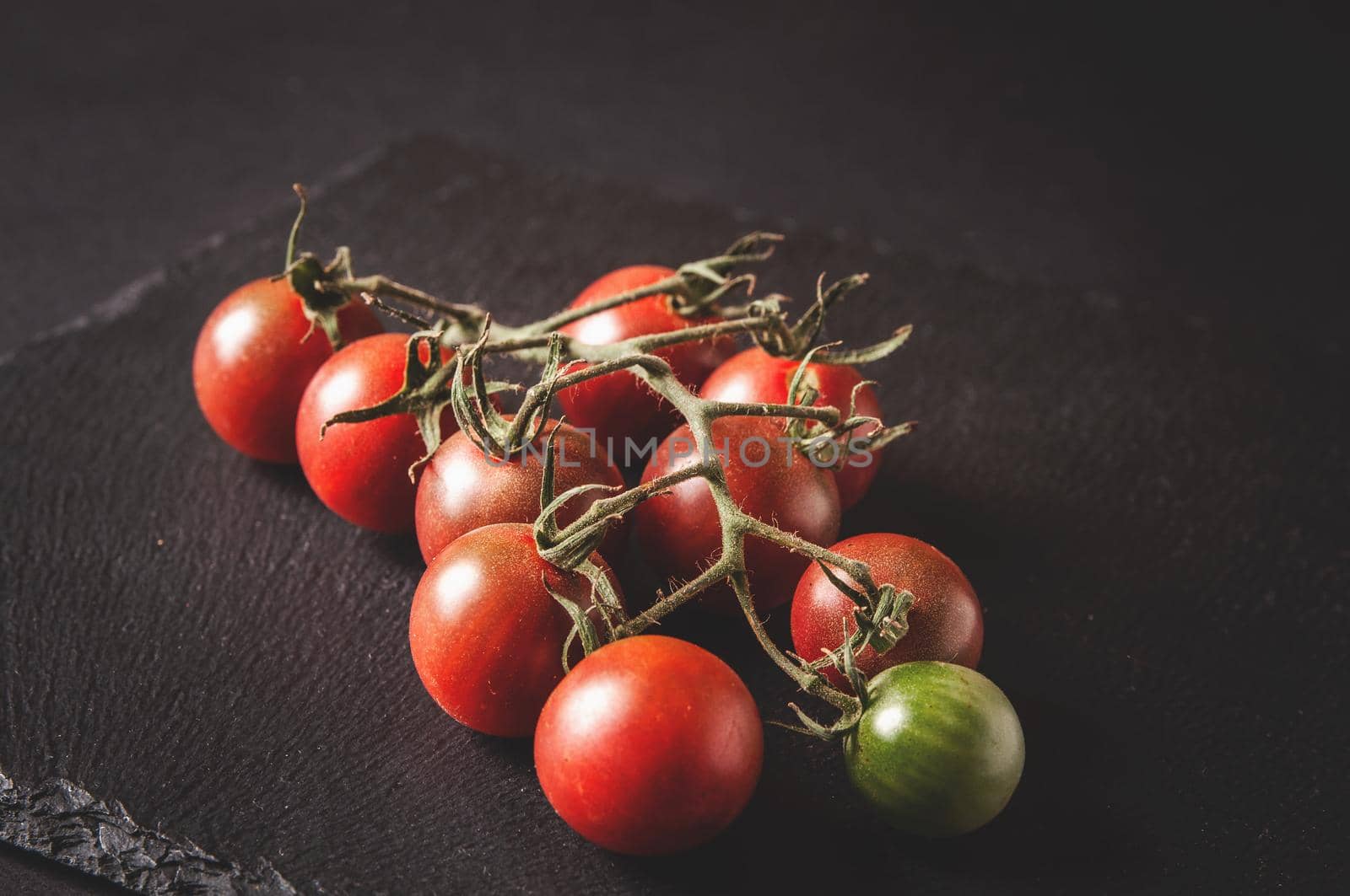 bunch of fresh cherry tomatoes on a branch are located on a black serving board on a dark background