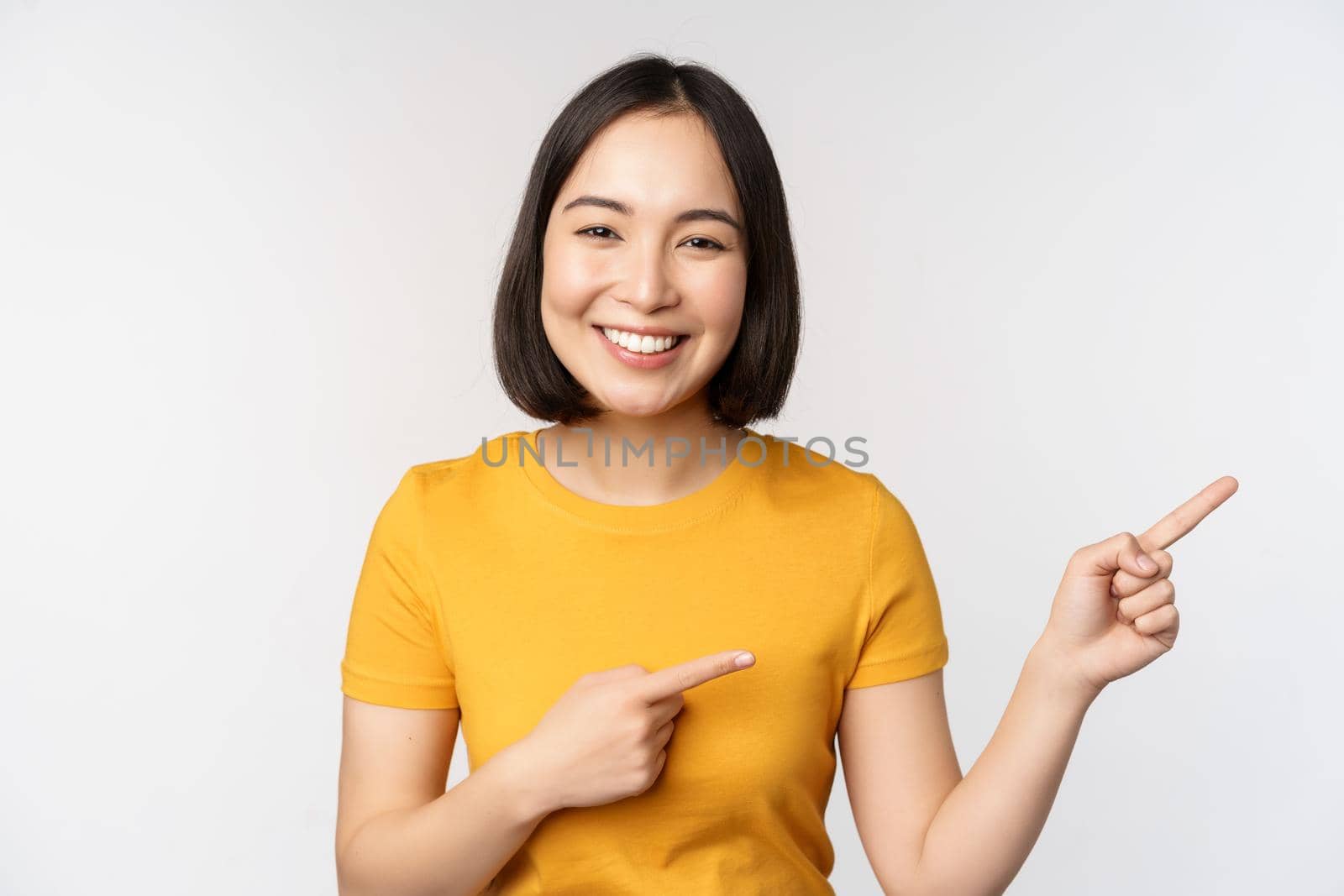 Amazed young asian woman, showing advertisement aside, pointing fingers right at promotion text, brand logo, standing happy against white background.