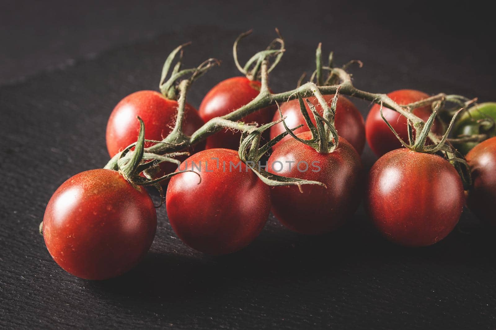 bunch of fresh cherry tomatoes on a branch are located on a black serving board on a dark background