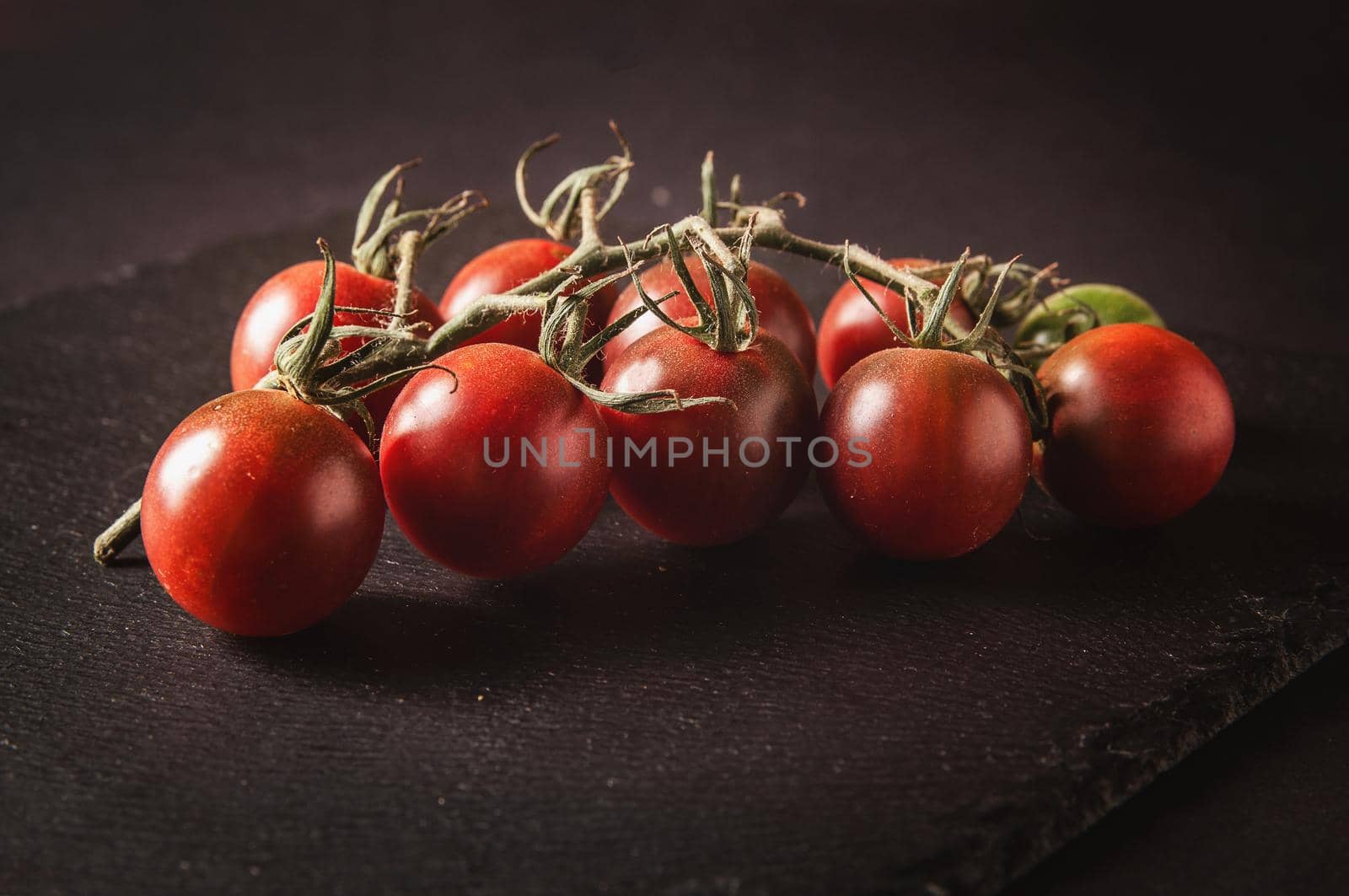 Bunch of fresh cherry tomatoes on a branch are located on a black serving board on a dark background