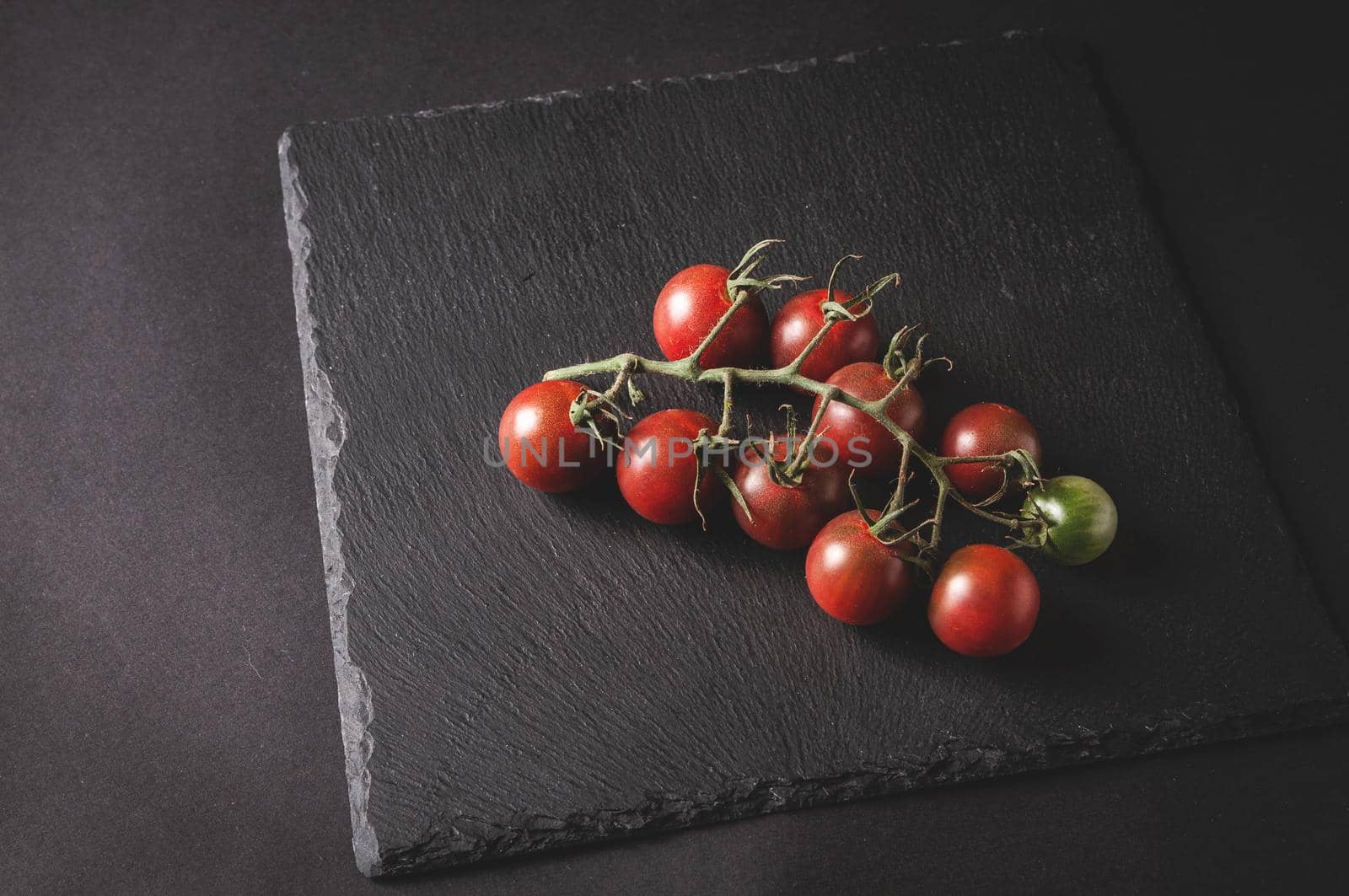 bunch of fresh cherry tomatoes on a branch are located on a black serving board by ozornina