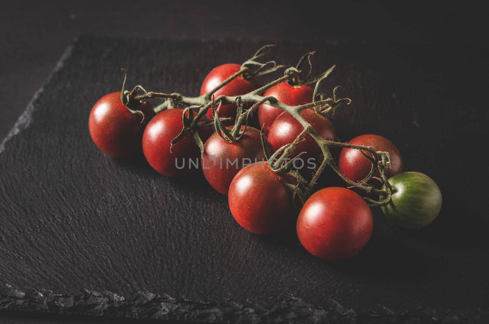 Bunch of fresh cherry tomatoes on a branch are located on a black serving board on a dark background