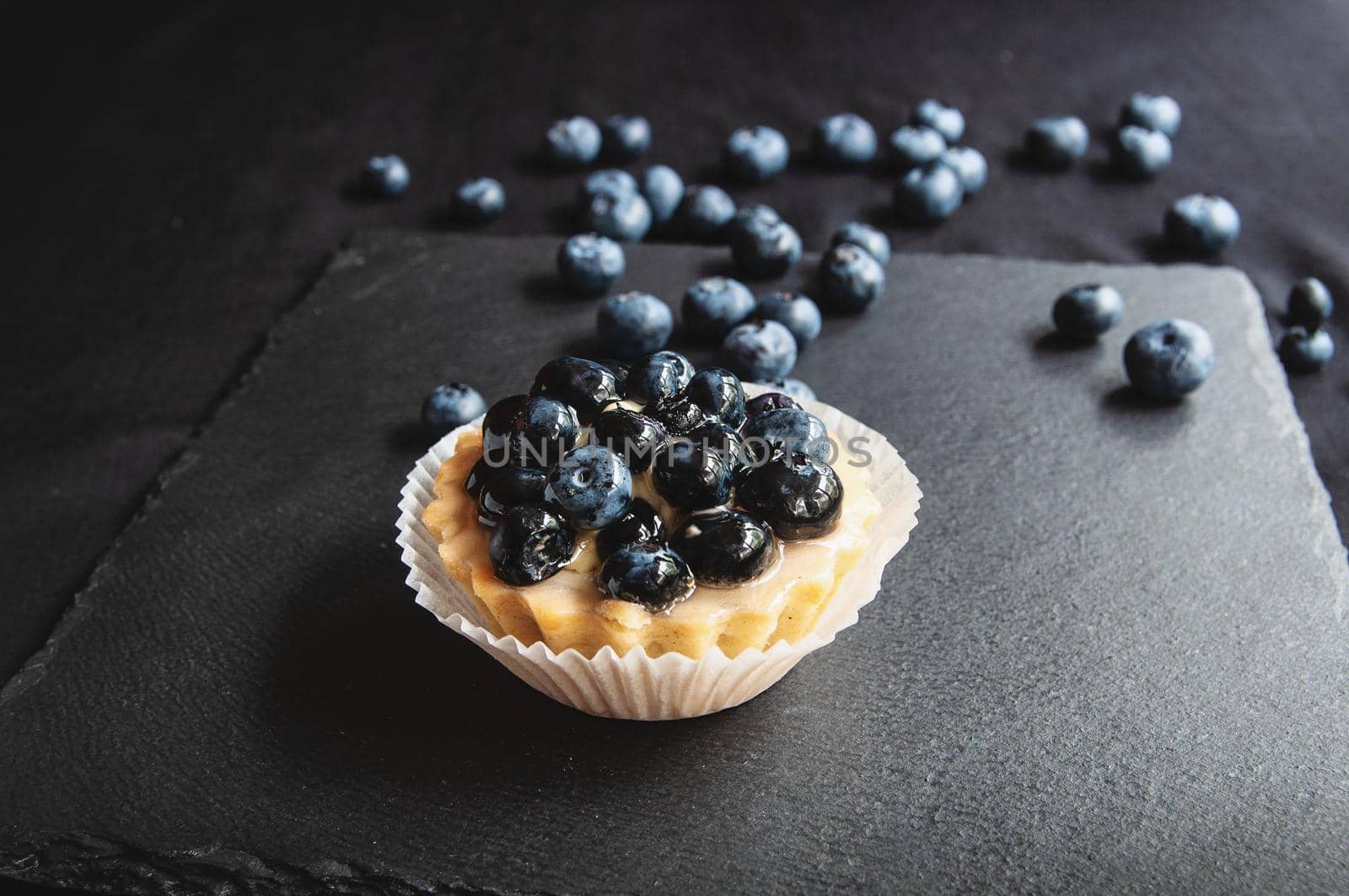 Delicious and sweet tarts made from cream and blueberry. Blueberry tart on a dark background. Blueberry cupcake on black serving board with scattered berries